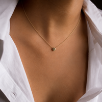 A delicate gold chain necklace with a small, round gold dot pendant worn by a model in an open white button-down shirt.
