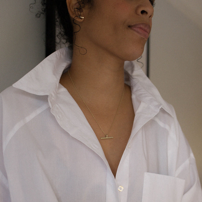 A close-up of a person wearing an oversized white button-up shirt with a solid gold T bar necklace on a Trace chain