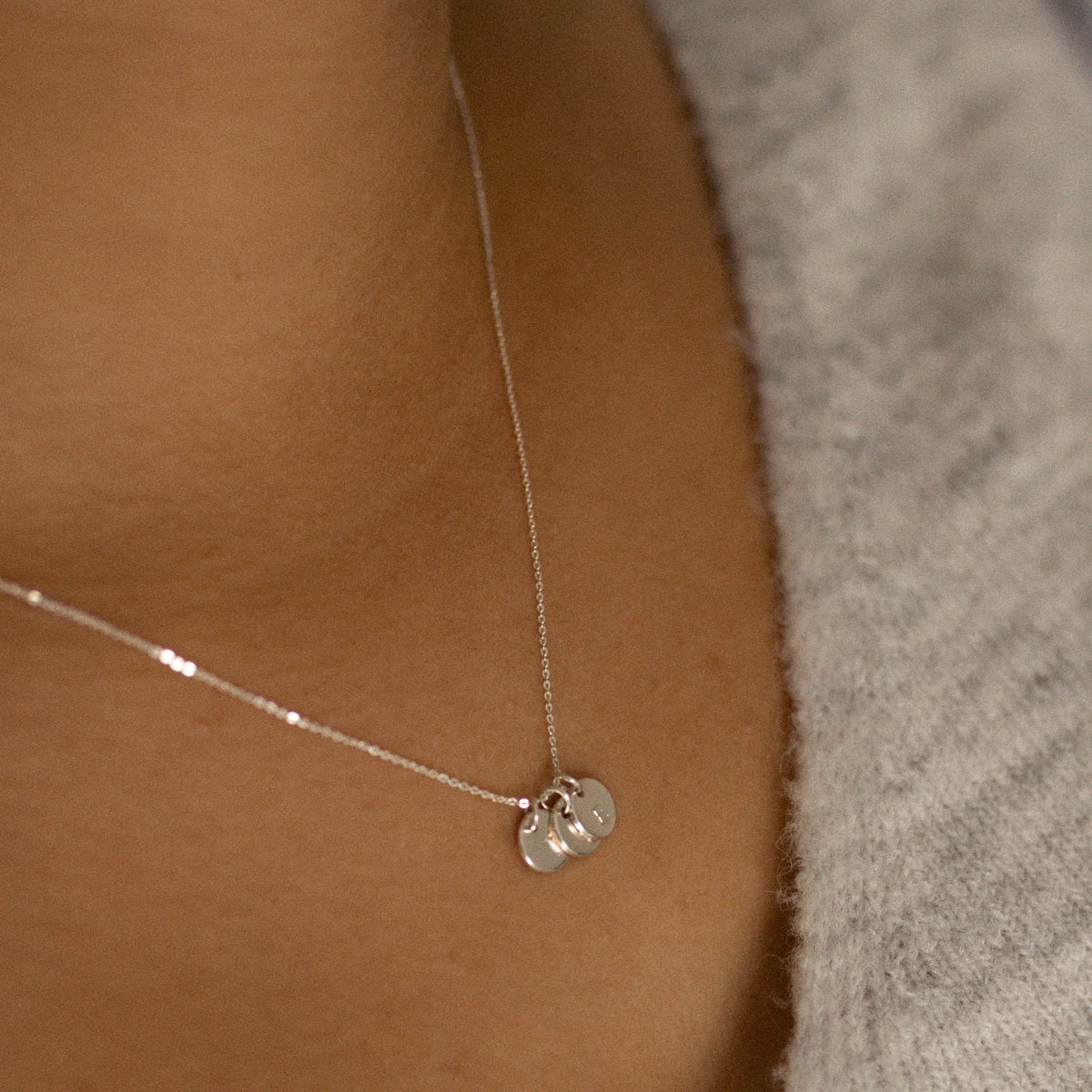 A close-up of a delicate 9ct white gold  chain with tiny discs personalised with an initial worn on a person's neck, against a gray knit top.