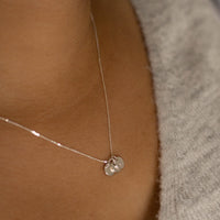 A close-up of a delicate 9ct white gold  chain with tiny discs personalised with an initial worn on a person's neck, against a gray knit top.